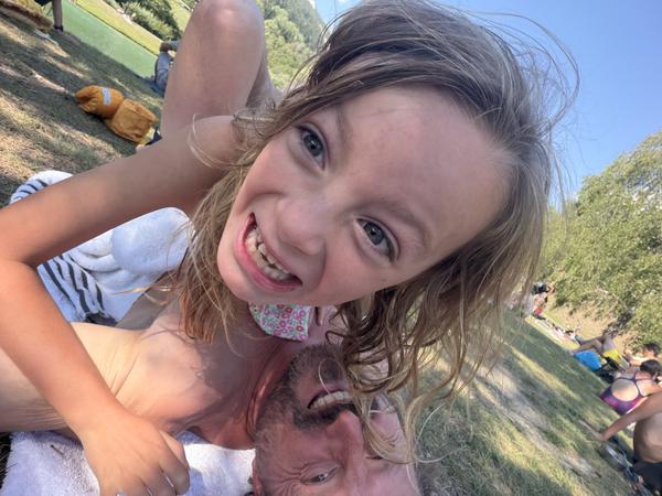 A child and an adult are captured in a joyful, playful selfie, lying on a sunny grassy bank at Rue du Lac, Bozel